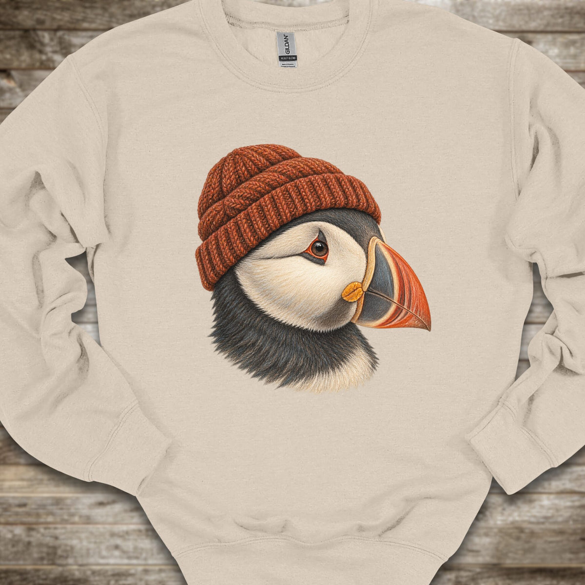 A cream-colored crewneck sweatshirt featuring a detailed graphic of a puffin wearing a rust-orange knit beanie, laid flat on a rustic wooden background.