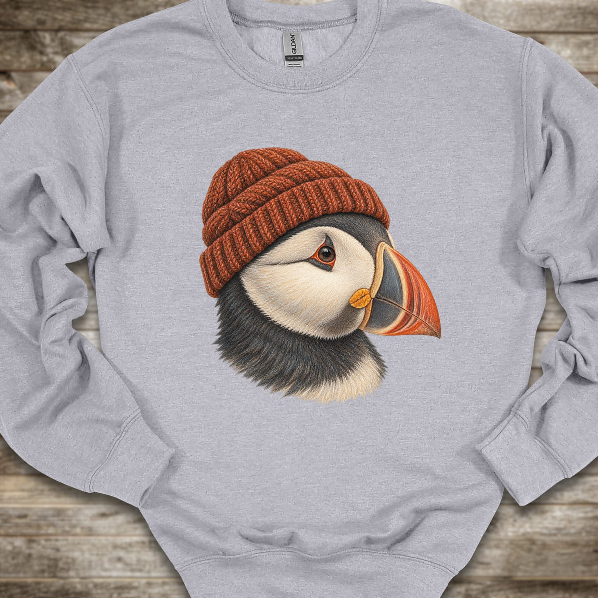 A light heather gray crewneck sweatshirt featuring a detailed graphic of a puffin wearing a brown knit beanie, laid flat on a rustic wooden surface.