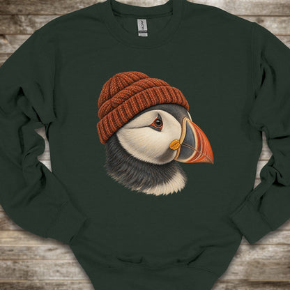 A dark forest green crewneck sweatshirt featuring a detailed graphic of a puffin wearing a rust-orange knit beanie, laid flat on a rustic wooden background.