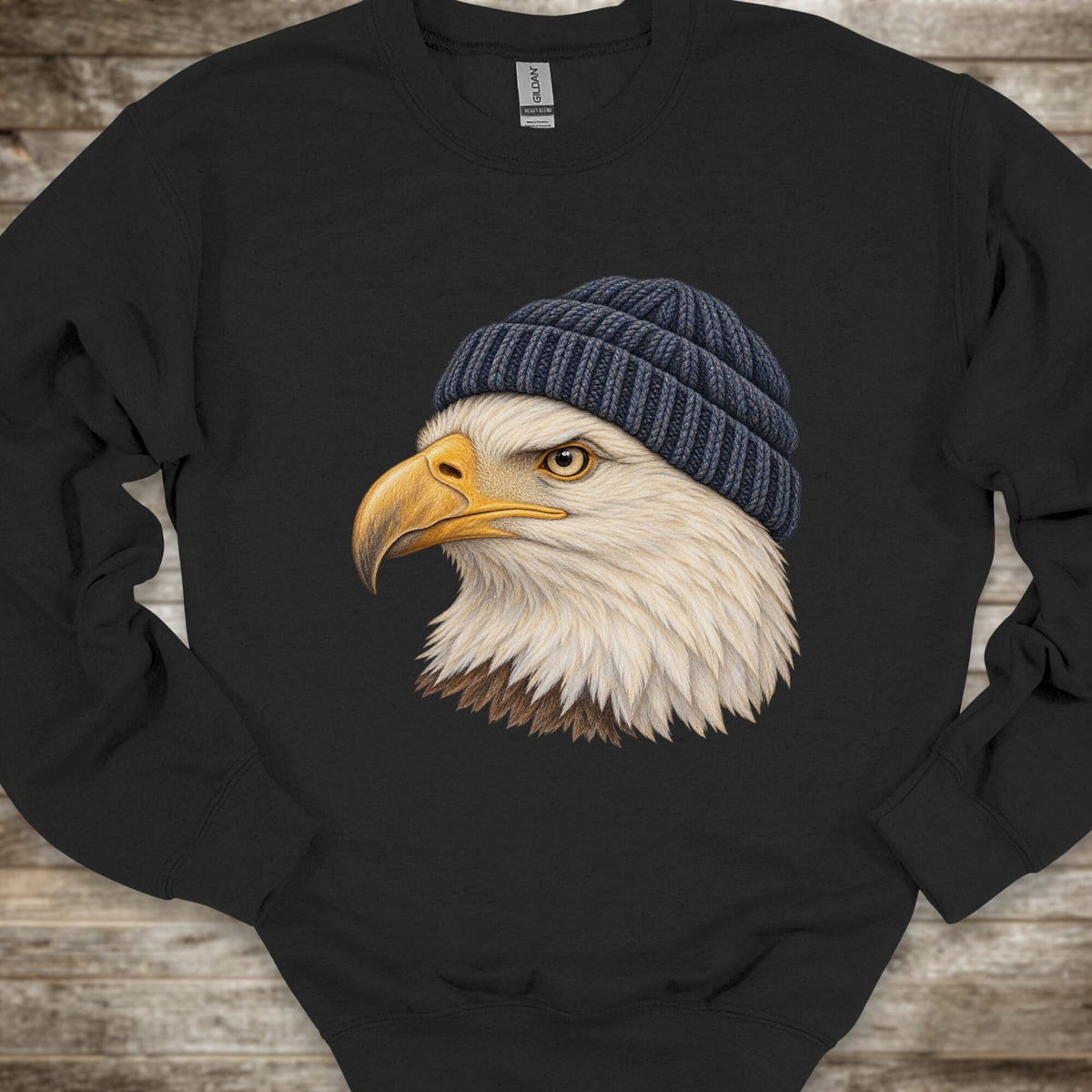 A black crewneck sweatshirt featuring a detailed graphic of a bald eagle's head wearing a dark blue knit beanie, displayed on a rustic wooden background.