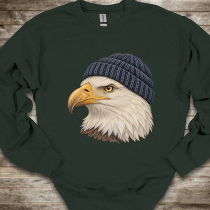 A dark forest green crewneck sweatshirt featuring a detailed graphic of a bald eagle wearing a navy blue knit beanie, laid flat on a rustic wooden background.