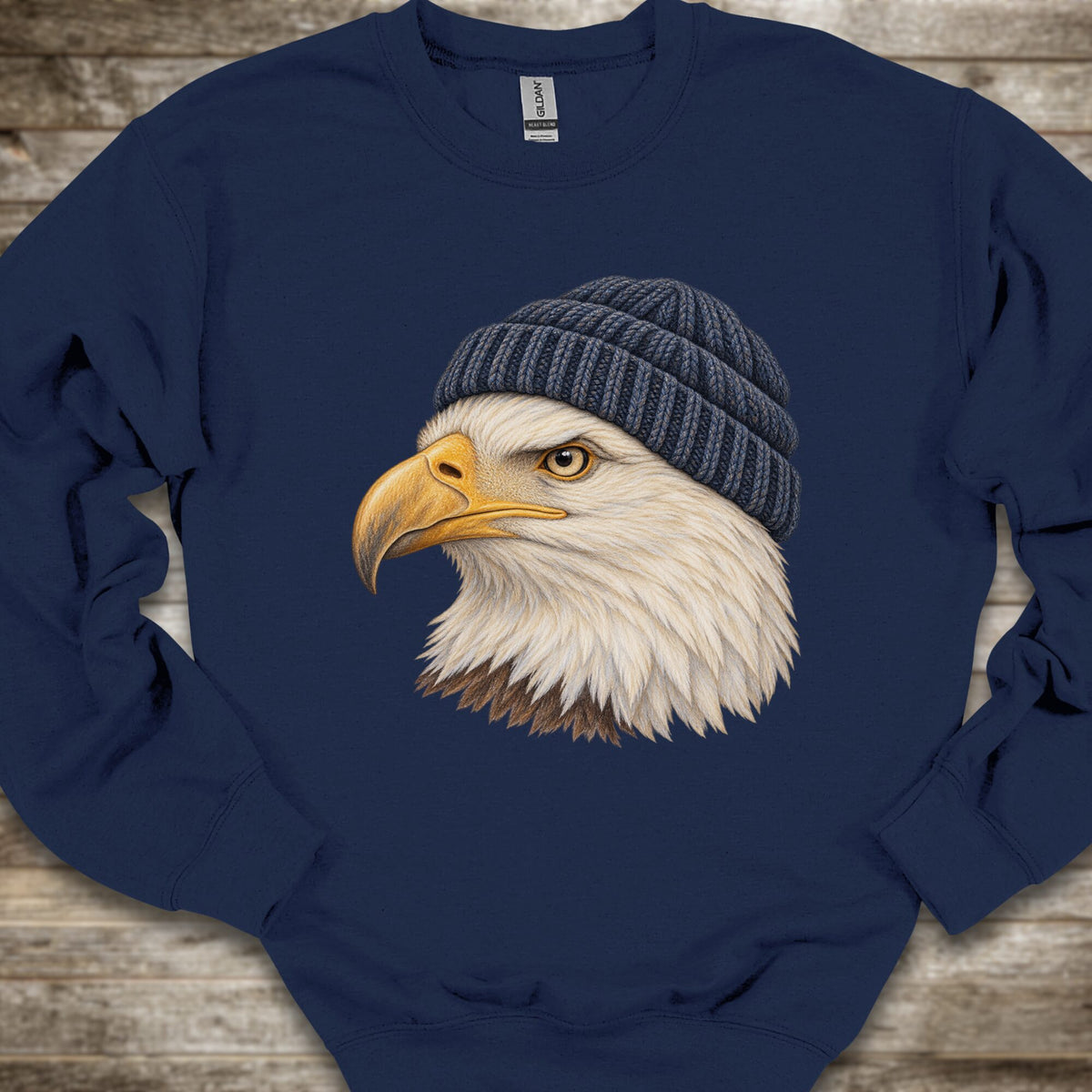 A navy blue crewneck sweatshirt featuring a detailed graphic of a bald eagle wearing a dark gray knit beanie, laid flat on a rustic wooden background.