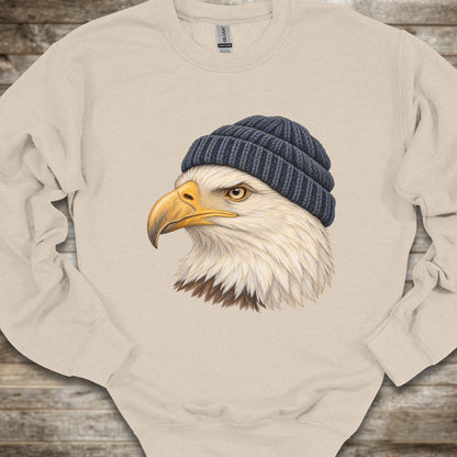 A beige crewneck sweatshirt featuring a graphic print of a bald eagle wearing a dark blue knit beanie, laid flat on a rustic wooden background.