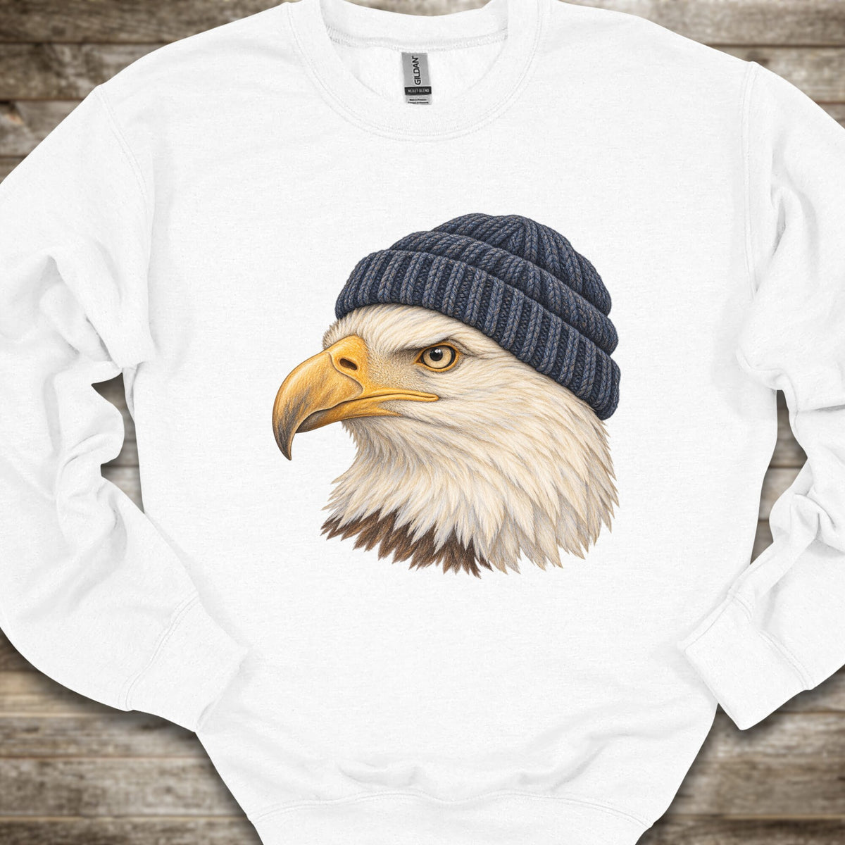 A white long-sleeve crewneck sweatshirt laid flat on a rustic wooden surface, featuring a detailed graphic of a bald eagle wearing a dark navy blue knit beanie on the front.