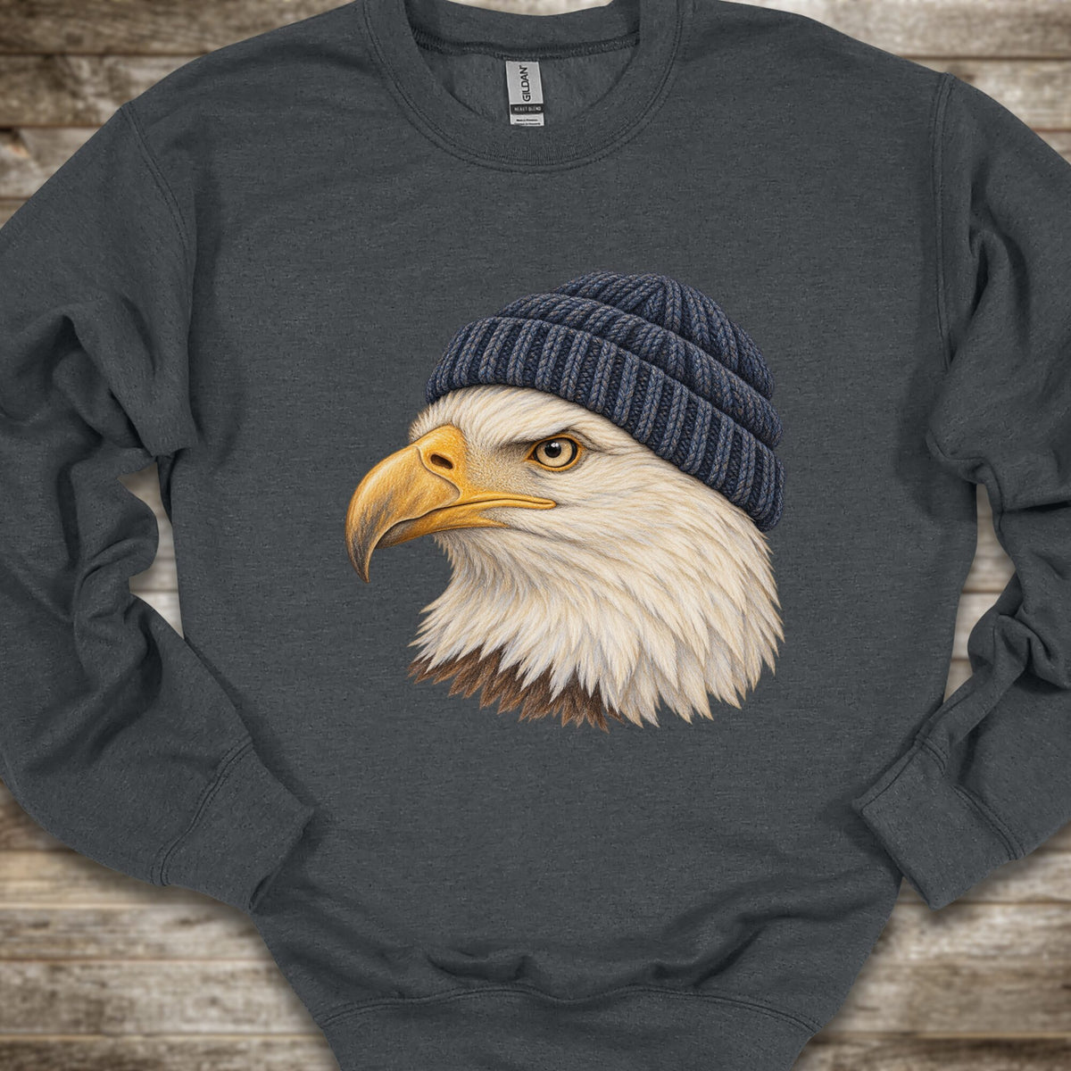 A dark gray crewneck sweatshirt featuring a graphic print of a bald eagle wearing a navy blue knit beanie, laid flat on a rustic wooden background.