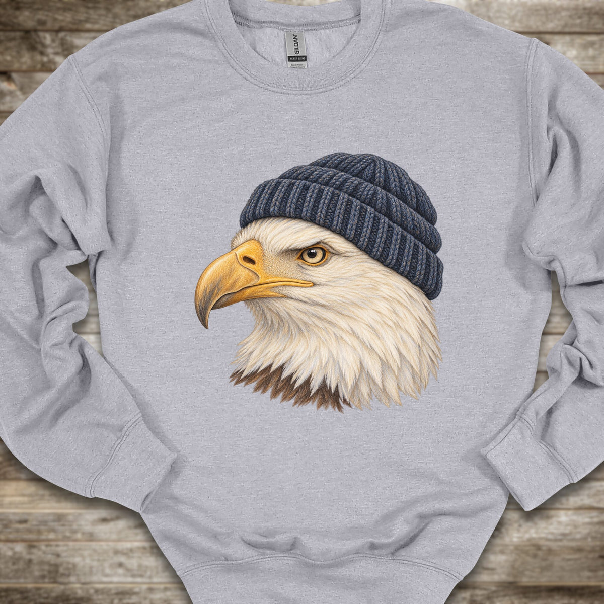 A light heather gray crewneck sweatshirt featuring a detailed graphic of a bald eagle's head wearing a dark navy blue knit beanie, laid flat on a rustic wooden surface.