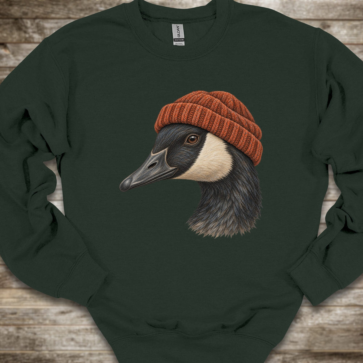A dark forest green crewneck sweatshirt featuring a detailed graphic of a goose wearing a rust-orange knit beanie, laid flat on a rustic wooden background.