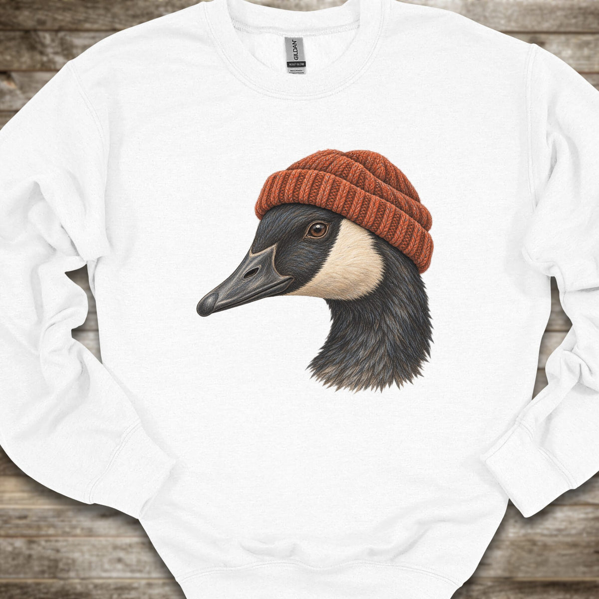 A white long-sleeve crewneck sweatshirt laid flat on a rustic wooden surface, featuring a graphic print of a goose wearing a rust-orange knit beanie.