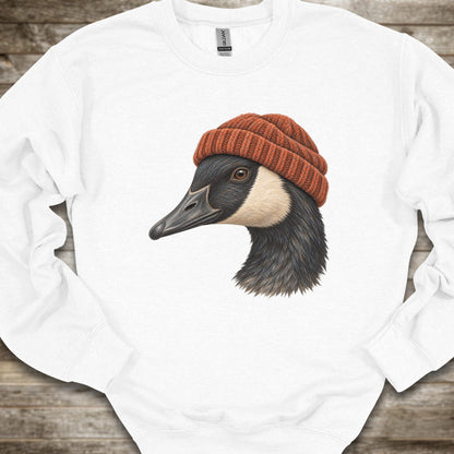 A white long-sleeve crewneck sweatshirt laid flat on a rustic wooden surface, featuring a graphic print of a goose wearing a rust-orange knit beanie.