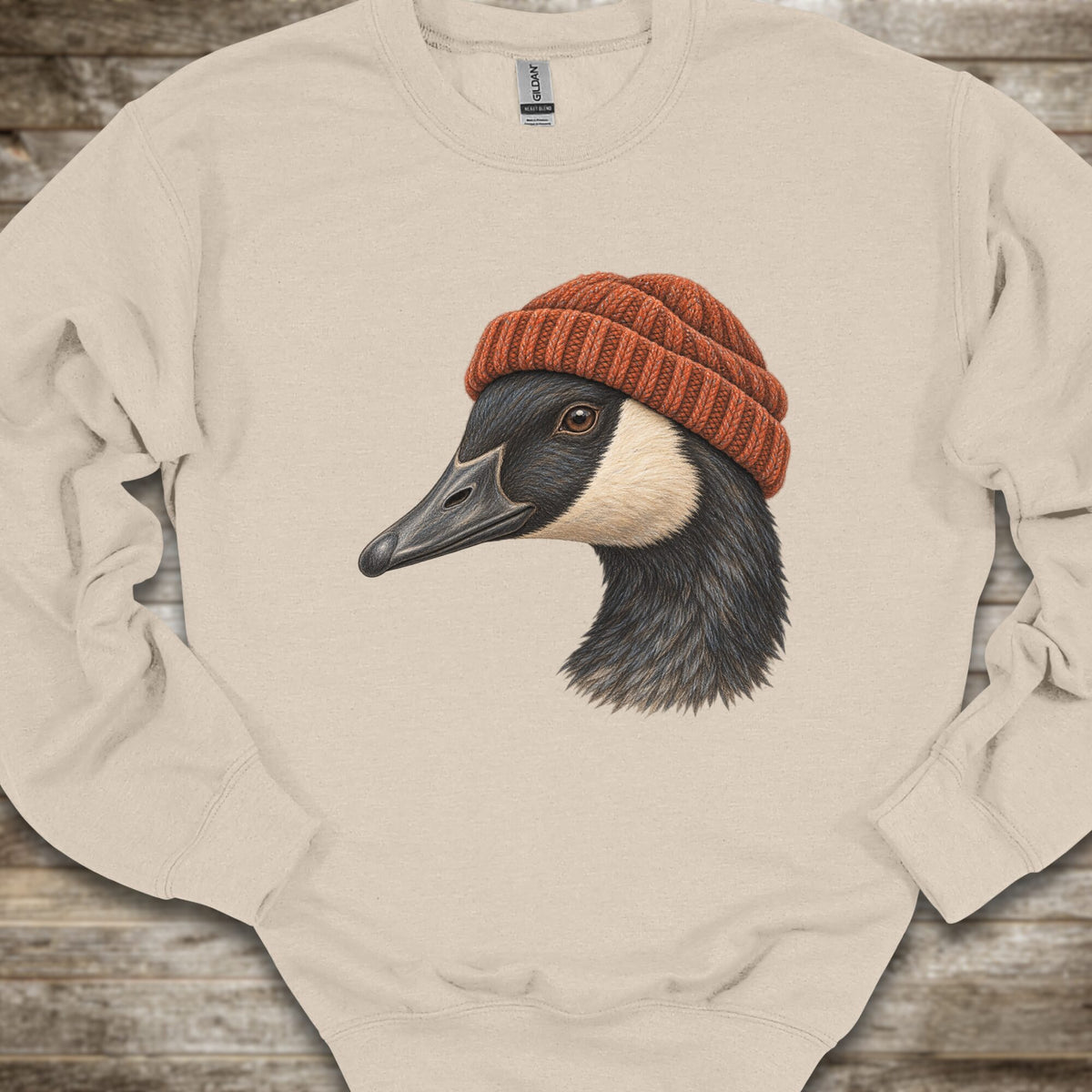 A beige crewneck sweatshirt laid flat on a rustic wooden surface, featuring a detailed graphic of a goose wearing a rust-orange knit beanie.