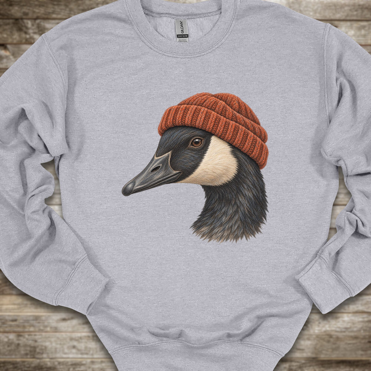 A light heather gray crewneck sweatshirt featuring a detailed graphic of a goose wearing a rust-orange knit beanie, laid flat on a rustic wooden surface.