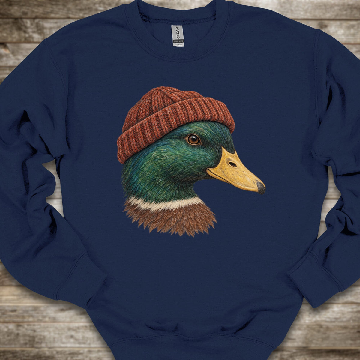 A navy blue crewneck sweatshirt featuring a graphic print of a mallard duck wearing a brown knit beanie, laid flat on a rustic wooden background.