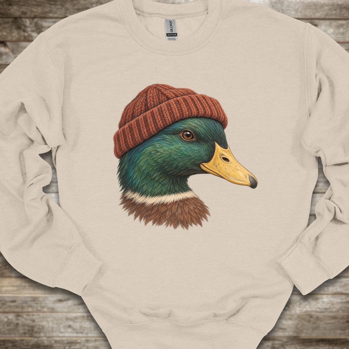 A cream-colored crewneck sweatshirt featuring a detailed graphic of a mallard duck wearing a rust-red knit beanie, laid flat on a rustic wooden surface.