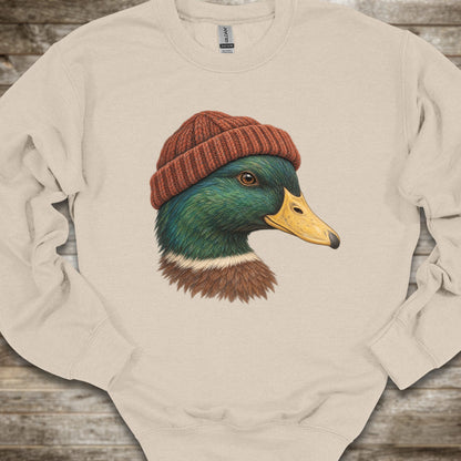 A cream-colored crewneck sweatshirt featuring a detailed graphic of a mallard duck wearing a rust-red knit beanie, laid flat on a rustic wooden surface.