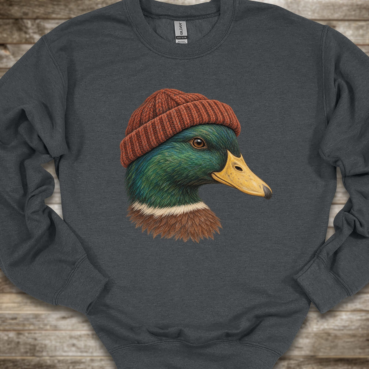 A dark gray crewneck sweatshirt featuring a graphic print of a mallard duck wearing a brown knit beanie, laid flat on a rustic wooden background.