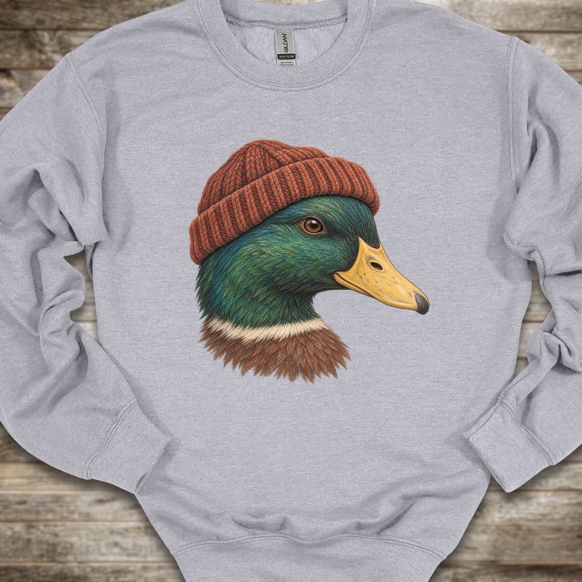 A light heather gray crewneck sweatshirt featuring a detailed graphic of a mallard duck wearing a rust-red knit beanie, laid flat on a rustic wooden surface.