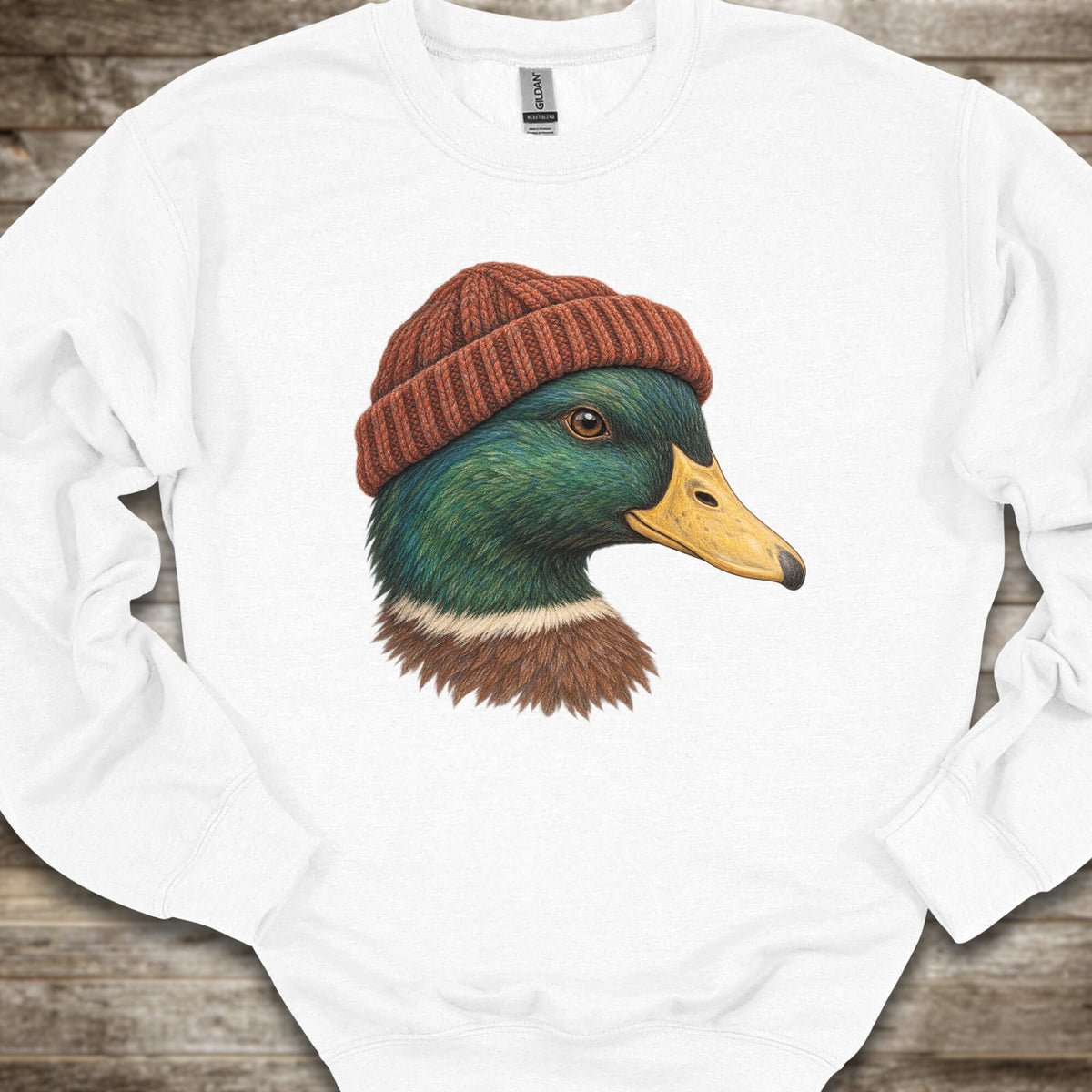 A white crewneck sweatshirt featuring a graphic print of a mallard duck wearing a brown knit beanie, laid flat on a rustic wooden background.