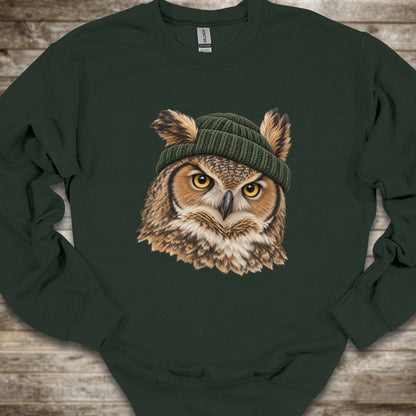 A dark forest green crewneck sweatshirt featuring a detailed graphic of a great horned owl with yellow eyes wearing a green knit beanie, laid flat on a rustic wooden background.