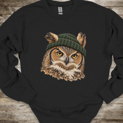 A black crewneck sweatshirt featuring a detailed graphic of a great horned owl with yellow eyes wearing a green knit beanie, laid flat on a rustic wooden background.