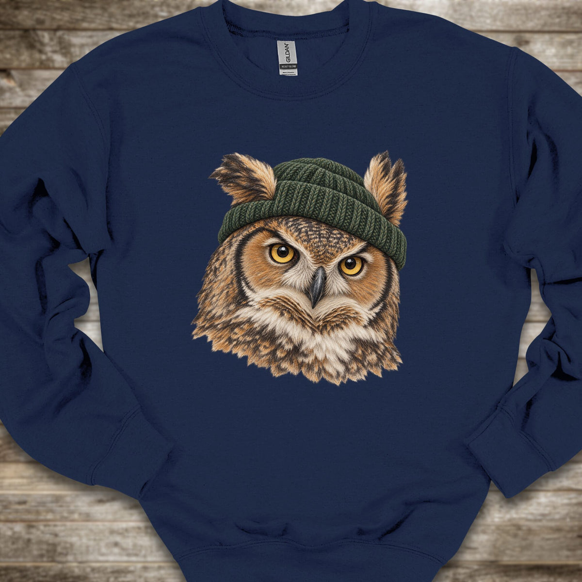 A navy blue crewneck sweatshirt featuring a detailed graphic of a brown owl with yellow eyes wearing a green knit beanie, laid flat on a rustic wooden background.