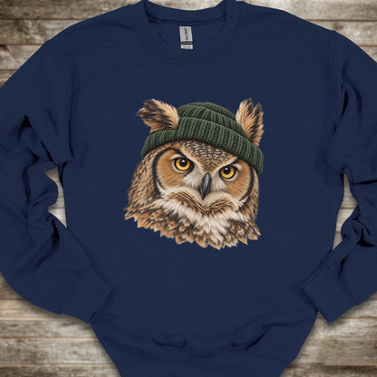 A navy blue crewneck sweatshirt featuring a detailed graphic of a brown owl with yellow eyes wearing a green knit beanie, laid flat on a rustic wooden background.
