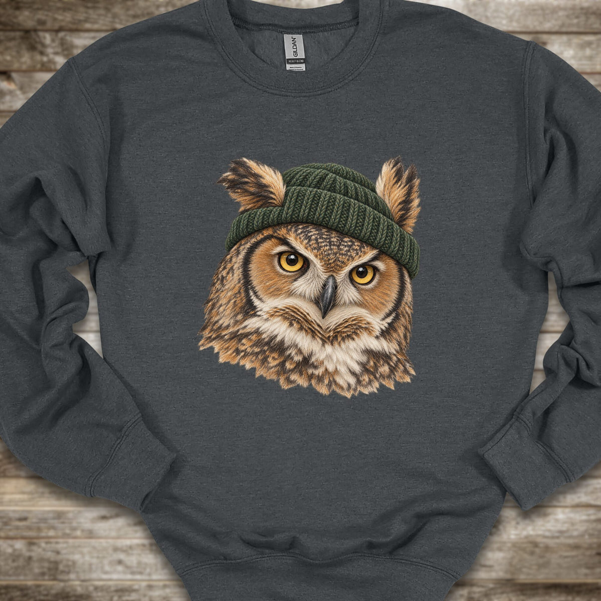 A dark gray crewneck sweatshirt featuring a graphic print of a great horned owl with yellow eyes wearing a green knit beanie, laid flat on a rustic wooden background.