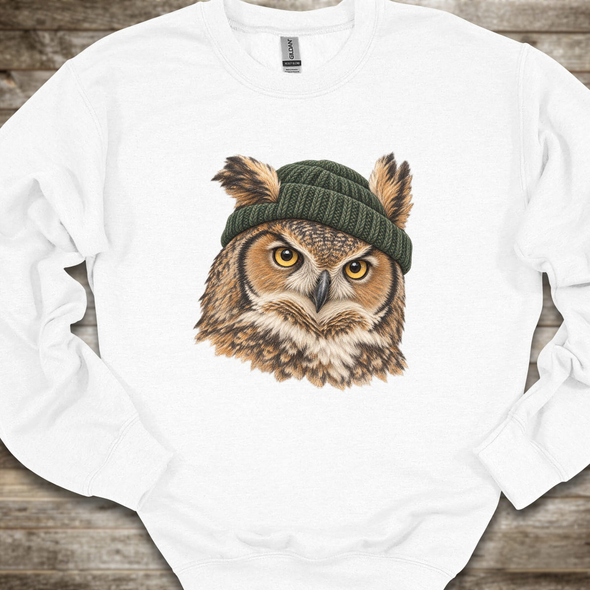 A white crewneck sweatshirt featuring a detailed graphic of a great horned owl with yellow eyes wearing a dark green knit beanie, laid flat on a rustic wooden background.