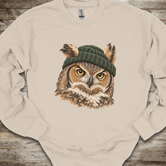 A cream-colored crewneck sweatshirt featuring a detailed graphic of a great horned owl with yellow eyes wearing a dark green knit beanie, laid flat on a rustic wooden background.