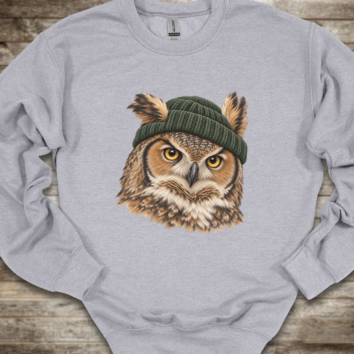 A light heather gray crewneck sweatshirt featuring a detailed graphic of a great horned owl with yellow eyes wearing a dark green knit beanie, laid flat on a rustic wooden surface.