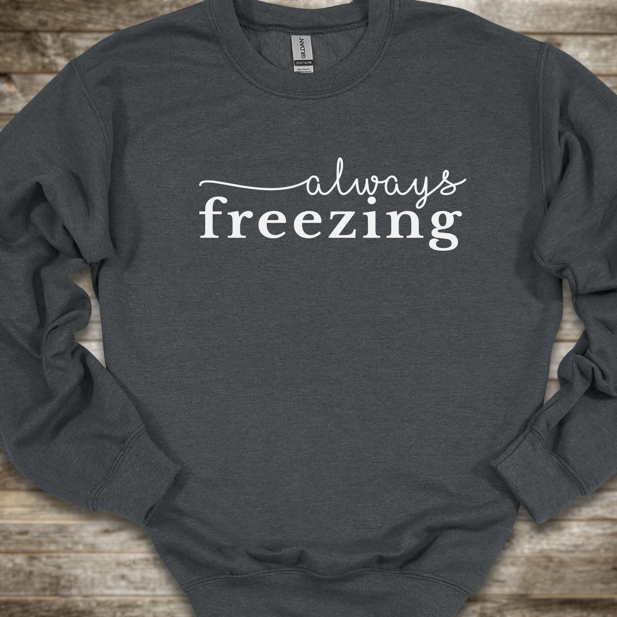 A dark gray heather crewneck sweatshirt with the phrase 'always freezing' printed in white across the chest, laid flat on a rustic wooden surface.
