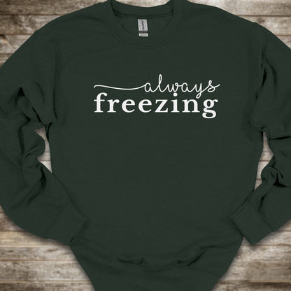 A dark forest green crewneck sweatshirt with the phrase 'always freezing' printed in white across the chest, laid flat on a rustic wooden background.