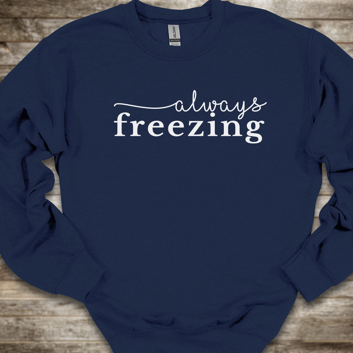 A navy blue crewneck sweatshirt with the phrase 'always freezing' printed in white across the chest, laid flat on a rustic wooden background.