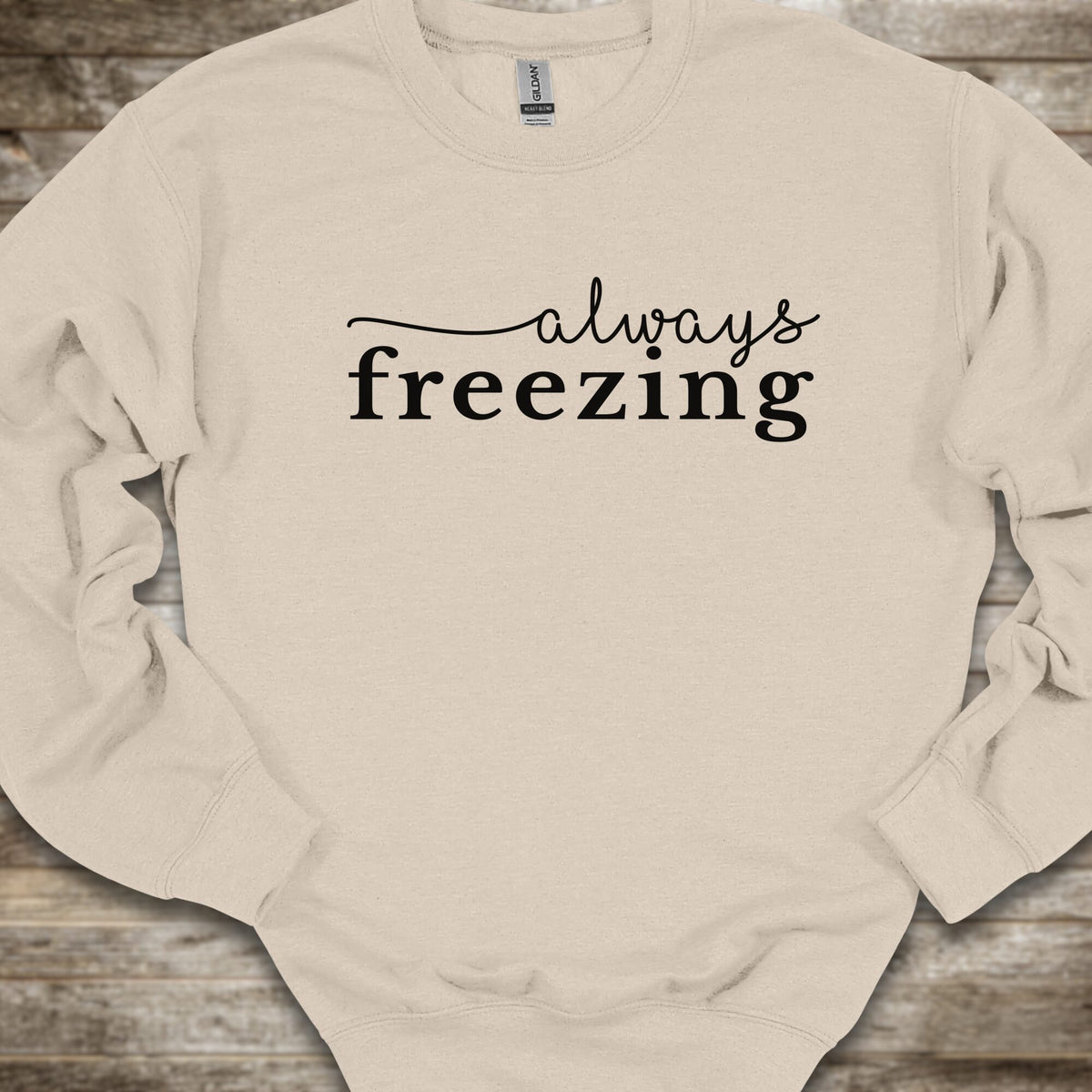 A light beige crewneck sweatshirt with the phrase 'always freezing' printed in black across the chest, laid flat on a rustic wooden surface.
