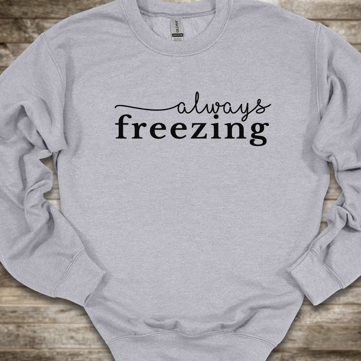 A light heather gray crewneck sweatshirt with the phrase 'always freezing' printed in black across the chest, laid flat on a rustic wooden surface.