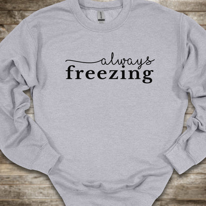 A light heather gray crewneck sweatshirt with the phrase 'always freezing' printed in black across the chest, laid flat on a rustic wooden surface.