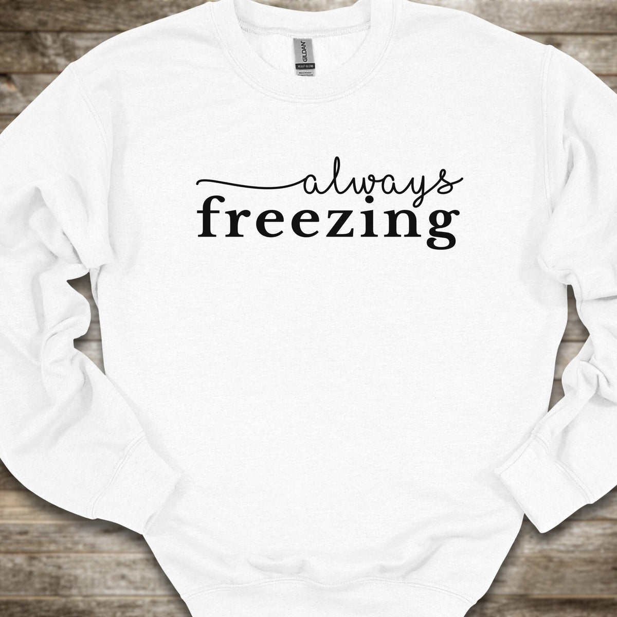 A white crewneck sweatshirt with the phrase 'always freezing' printed in black across the chest, laid flat on a rustic wooden background.