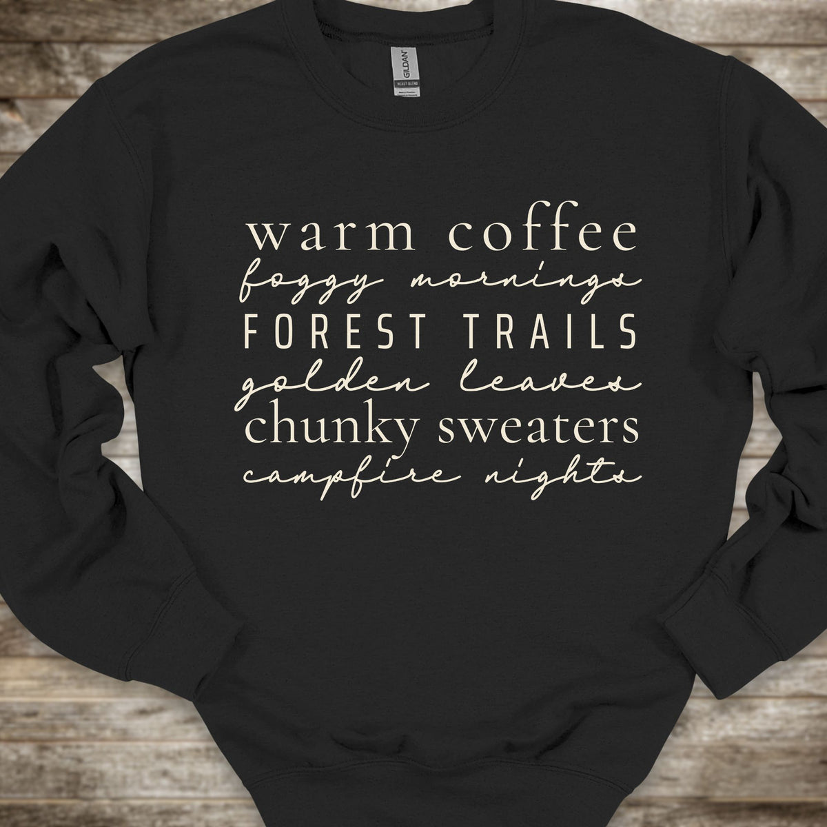 A black crewneck sweatshirt with a white typographic print listing cozy phrases like 'warm coffee', 'foggy morning', 'FOREST TRAILS', 'golden leaves', 'chunky sweaters', and 'campfire nights'. The sweatshirt is laid flat on a rustic wooden background.