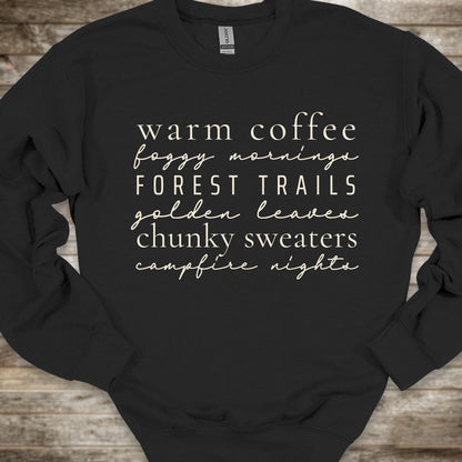 A black crewneck sweatshirt with a white typographic print listing cozy phrases like 'warm coffee', 'foggy morning', 'FOREST TRAILS', 'golden leaves', 'chunky sweaters', and 'campfire nights'. The sweatshirt is laid flat on a rustic wooden background.