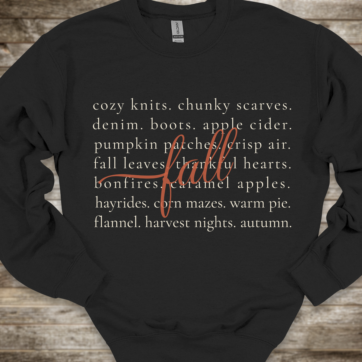 A black crewneck sweatshirt with a cozy autumn-themed text graphic on the front, laid flat on a rustic wooden background. The graphic features a list of fall elements in a light beige font, overlaid with the word 'fall' in a cursive orange script.