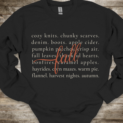 A black crewneck sweatshirt with a cozy autumn-themed text graphic on the front, laid flat on a rustic wooden background. The graphic features a list of fall elements in a light beige font, overlaid with the word 'fall' in a cursive orange script.