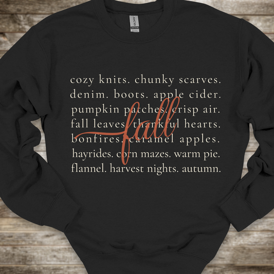 A black crewneck sweatshirt with a cozy autumn-themed text graphic on the front, laid flat on a rustic wooden background. The graphic features a list of fall elements in a light beige font, overlaid with the word 'fall' in a cursive orange script.