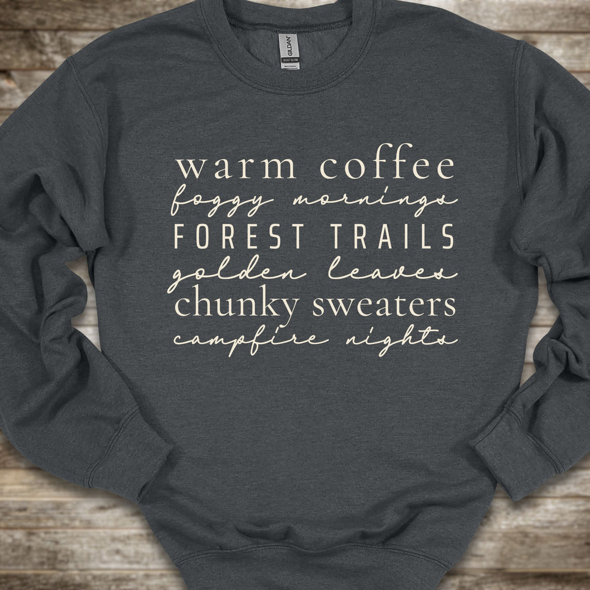 A dark charcoal gray crewneck sweatshirt with cream-colored text listing cozy phrases like 'warm coffee', 'foggy mornings', 'FOREST TRAILS', 'golden leaves', 'chunky sweaters', and 'campfire nights', laid flat on a rustic wooden background.