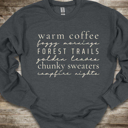 A dark charcoal gray crewneck sweatshirt with cream-colored text listing cozy phrases like 'warm coffee', 'foggy mornings', 'FOREST TRAILS', 'golden leaves', 'chunky sweaters', and 'campfire nights', laid flat on a rustic wooden background.