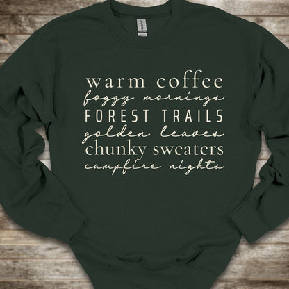 A dark forest green crewneck sweatshirt with white text listing cozy phrases like 'warm coffee', 'foggy mornings', 'FOREST TRAILS', 'golden leaves', 'chunky sweaters', and 'campfire nights', laid flat on a rustic wooden background.