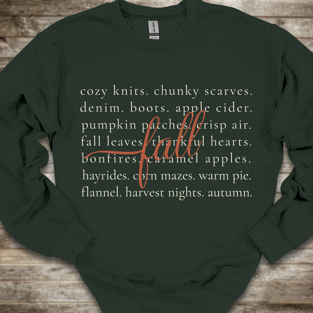 A dark forest green crewneck sweatshirt with a cozy autumn-themed text graphic on the front, laid flat on a rustic wooden background.