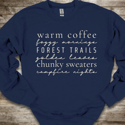 A navy blue crewneck sweatshirt with white text listing cozy phrases like 'warm coffee', 'foggy mornings', 'FOREST TRAILS', 'golden leaves', 'chunky sweaters', and 'campfire nights', laid flat on a rustic wooden background.