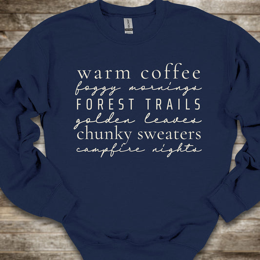 A navy blue crewneck sweatshirt with white text listing cozy phrases like 'warm coffee', 'foggy mornings', 'FOREST TRAILS', 'golden leaves', 'chunky sweaters', and 'campfire nights', laid flat on a rustic wooden background.