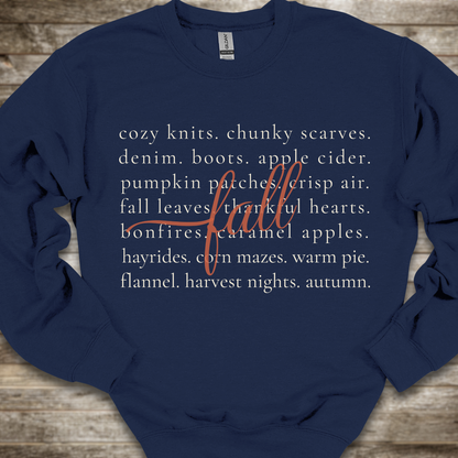 A navy blue crewneck sweatshirt with a white typographic print listing autumn-themed words and a large cursive word 'fall' in red, laid flat on a rustic wooden background.