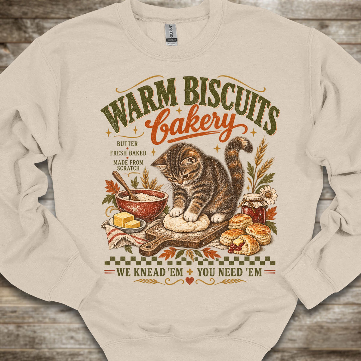 A beige crewneck sweatshirt featuring a vintage-style graphic of a tabby cat baking warm biscuits at a bakery. The graphic includes text reading 'WARM BISCUITS bakery' and 'WE KNEAD 'EM + YOU NEED 'EM', along with illustrations of a bowl of batter, butter, and baked goods, displayed on a rustic wooden background.