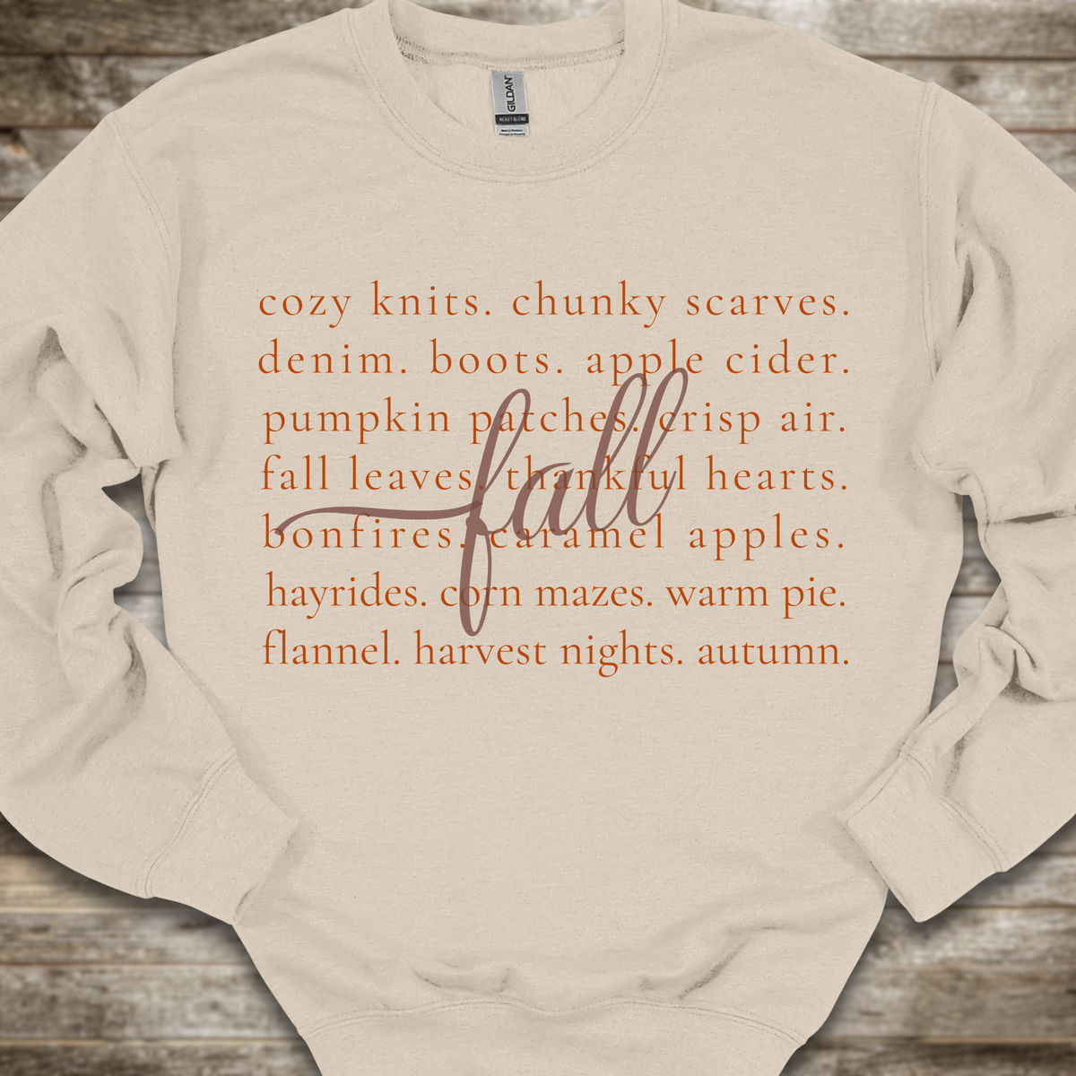 A cream-colored crewneck sweatshirt with a textured orange typographic print listing autumn themes and words, laid flat on a rustic wooden background.